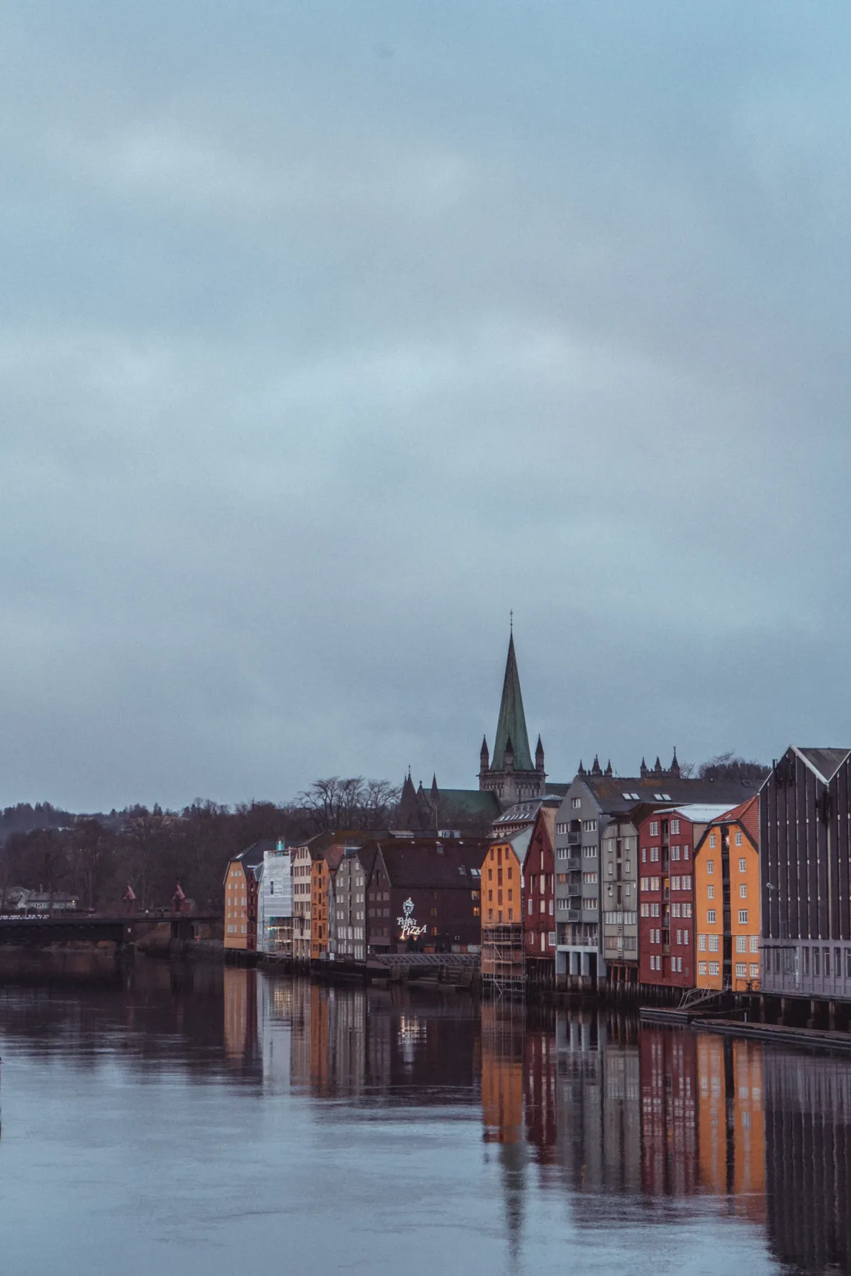 Colourful wooden warehouses and a church spire reflected in a calm river under a grey, overcast sky.