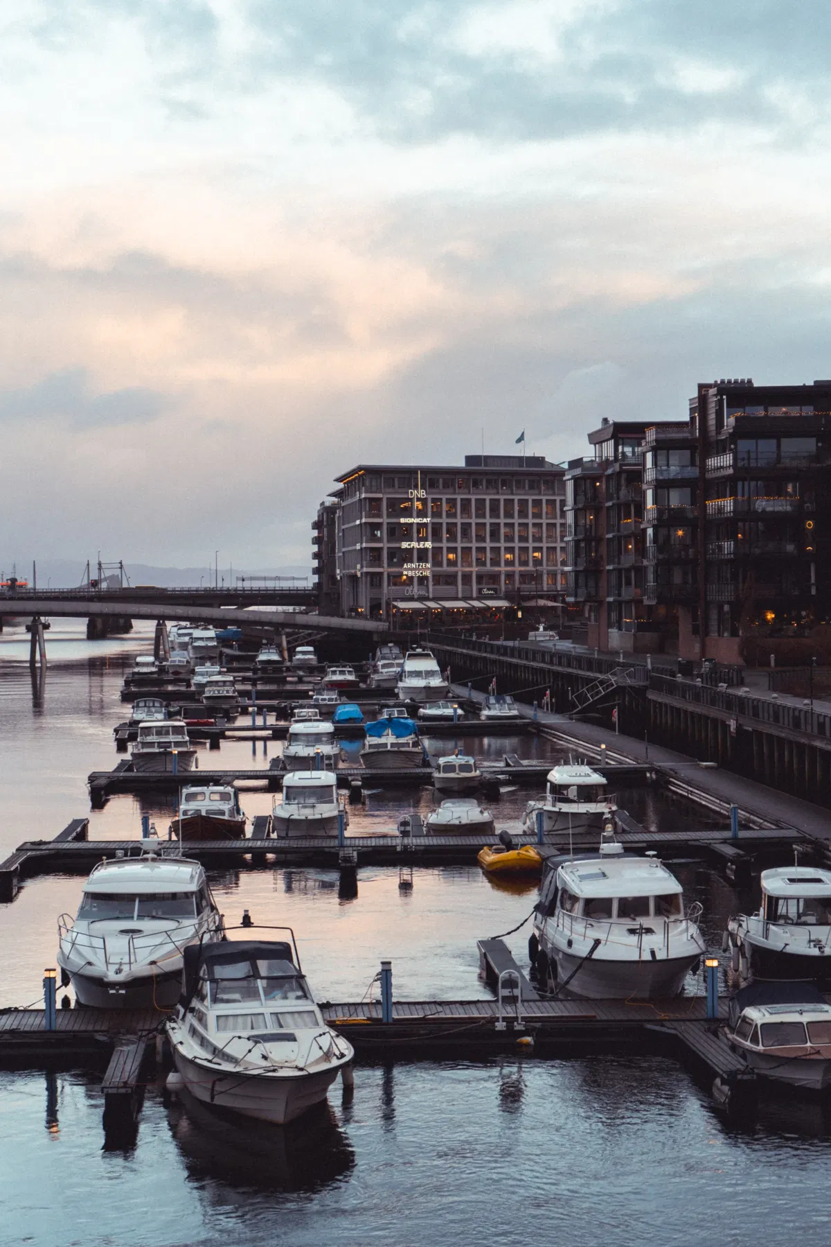 Motorboats moored at wooden docks in a harbour, with modern waterfront buildings and a bridge in soft dusk light.