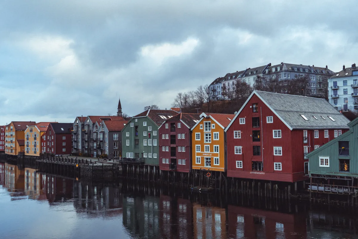 Red, yellow and green wooden warehouses on stilts along a calm river, with hillside apartment buildings under heavy clouds.
