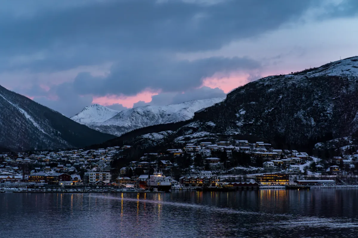 A snowy harbour town with lit windows beneath mountains and pink-tinged clouds at dusk, a ferry at the quay.