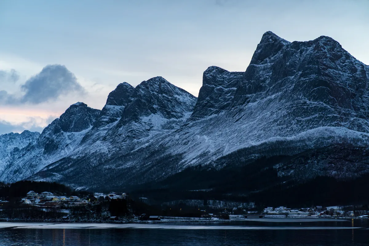 Jagged snow-dusted mountain ridges rising above a dark fjord, with a few houses on the shore in blue evening light.