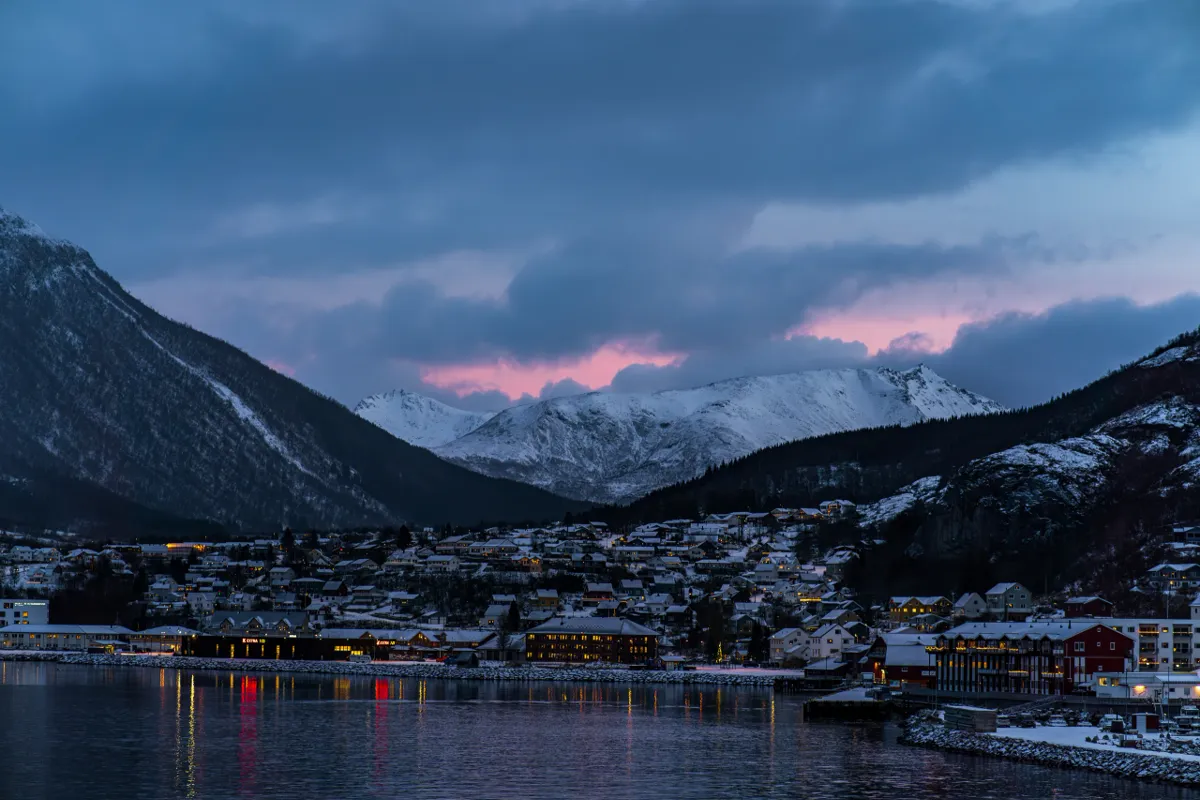 A fjord-side town with lights reflected in the water, below snowy mountains and a pink gap in dark clouds.