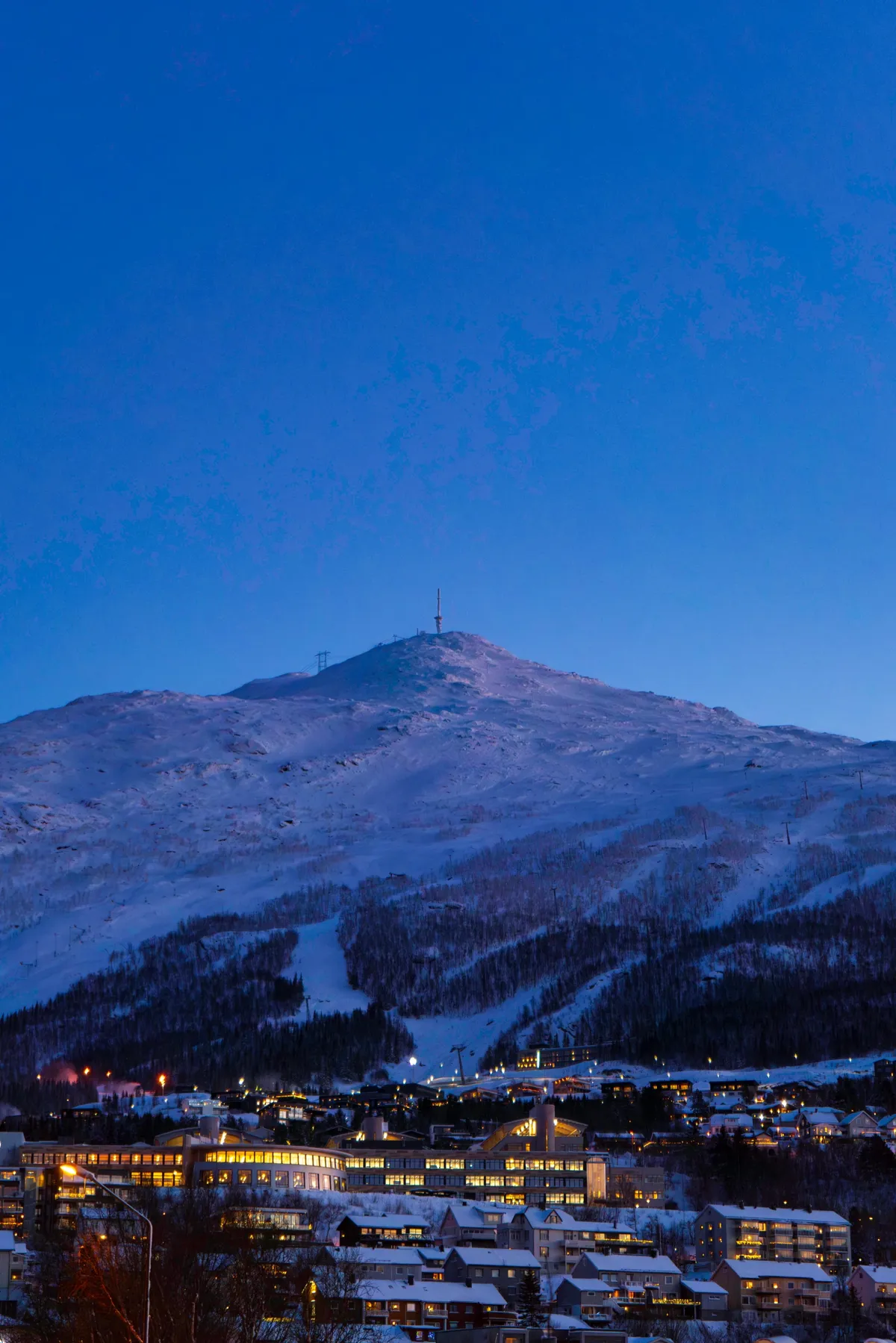 A snow-covered mountain with a mast on its summit above the lit town of Narvik at deep blue dusk.
