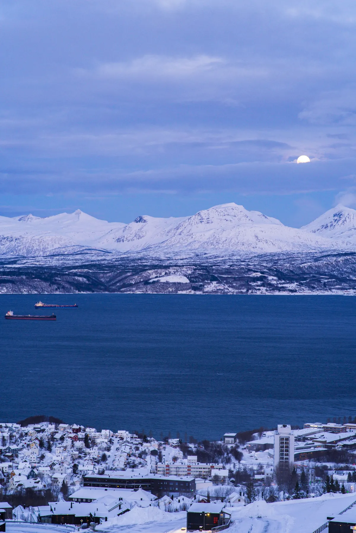 The moon rises over a dark blue fjord and snowy mountains, with cargo ships on the water and snowy Narvik.