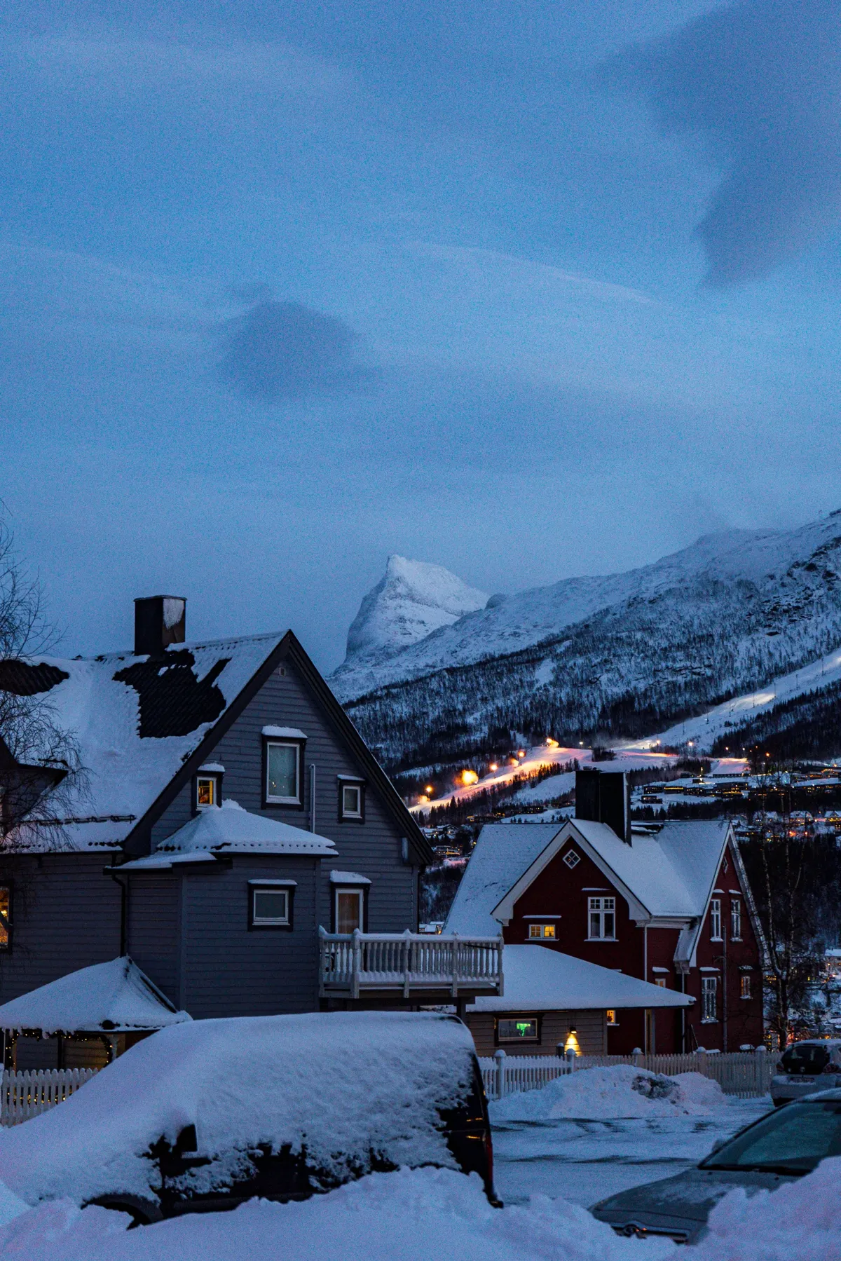 Snow-covered houses and parked cars in Narvik at dusk, with a pointed snowy peak and lit ski slope behind.