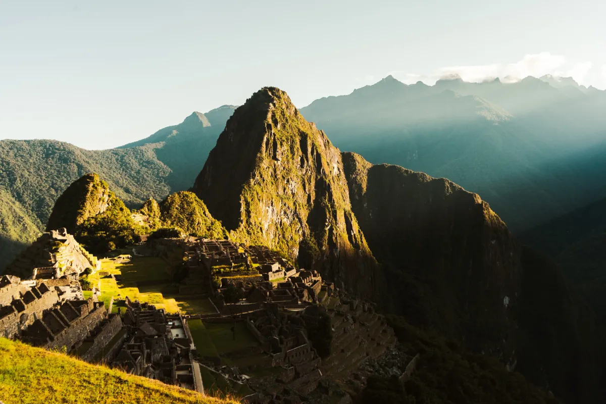 Early sun glances across the Machu Picchu ruins and the steep green peak behind, with mountains fading into haze.