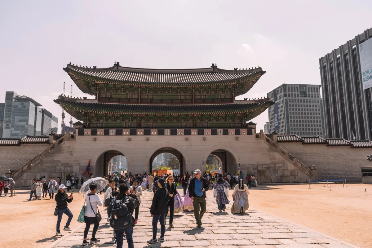 Two-storey palace gate with a tiled roof above three stone arches in Seoul, visitors in hanbok crossing the paved forecourt, modern towers behind under a hazy sky.