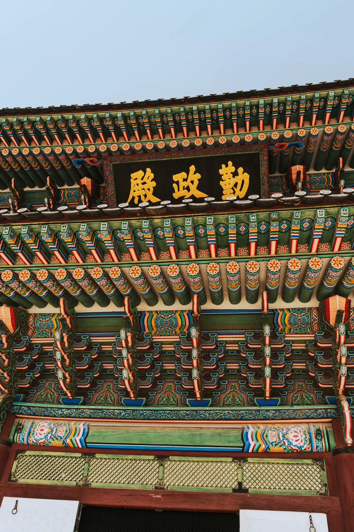 Underside of a palace roof in Seoul painted in green, red and blue, with a dark signboard of gold Chinese characters under a pale blue sky.