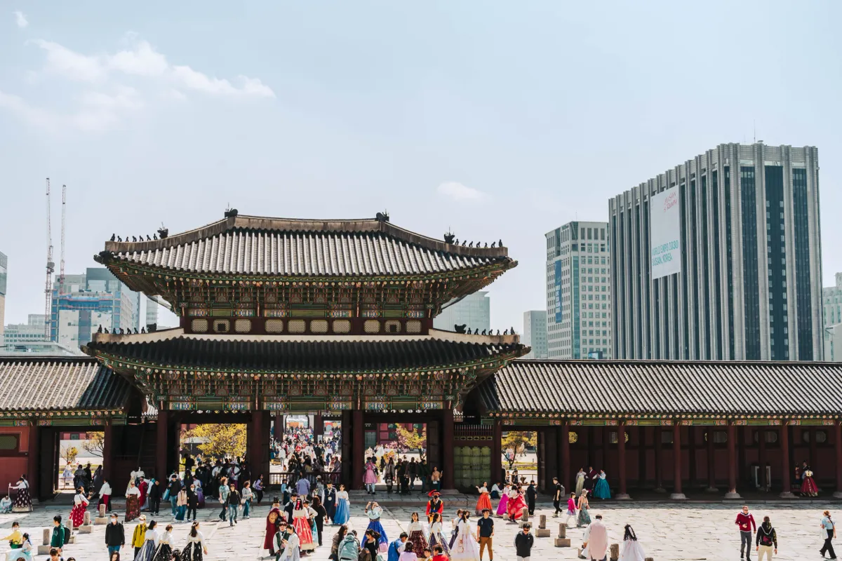 Palace gate with a grey tiled roof in Seoul, crowds in colourful hanbok in the courtyard in front, glass skyscrapers rising behind under a hazy blue sky.