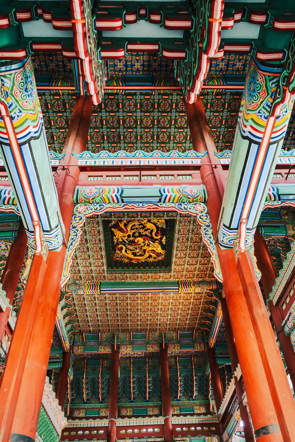 Looking up inside a palace hall in Seoul at red pillars and an ornately painted ceiling in green, blue and gold, with a golden dragon panel at the centre.