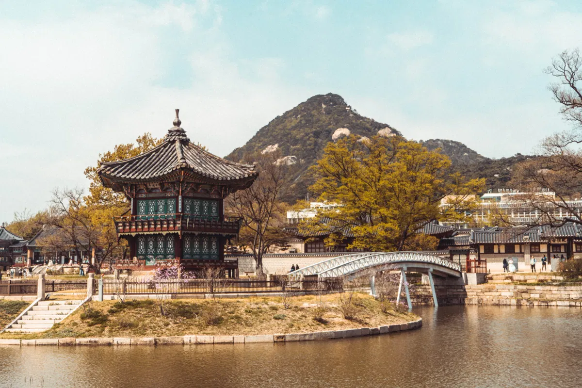 Hexagonal pavilion with a dark tiled roof on a small island in a pond, an arched white bridge beside it and a rocky mountain behind, in Seoul.