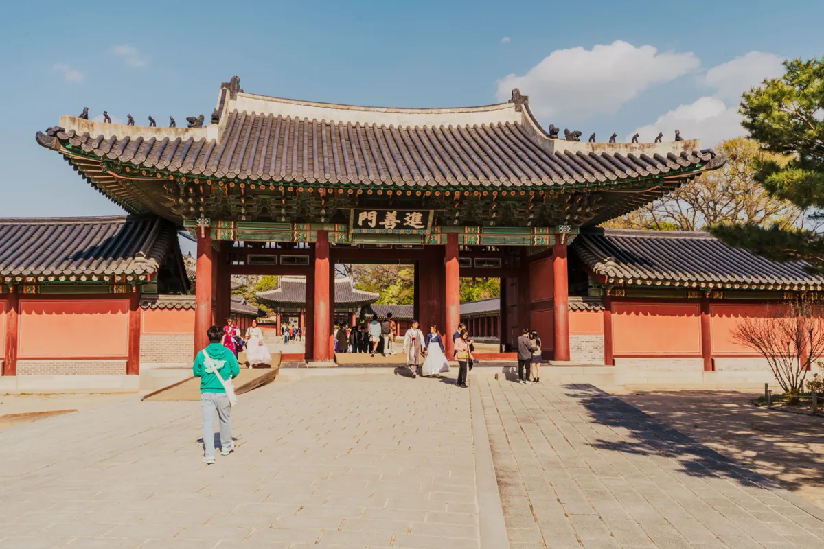 Palace gate with red pillars, orange walls and a dark tiled roof under a blue sky with white clouds, a few visitors crossing the sunlit stone courtyard.