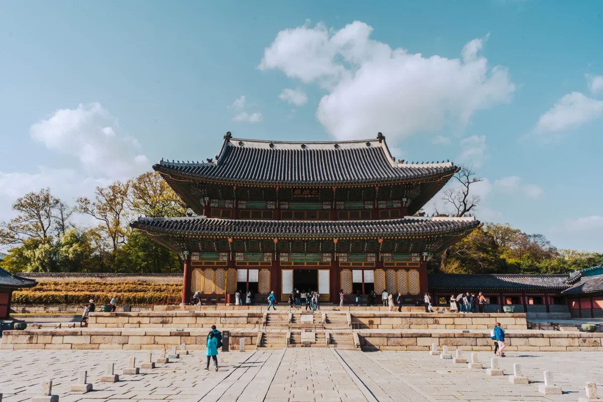 Palace hall with a two-tier dark tiled roof and red columns at the far end of a wide stone courtyard, under a blue sky with white clouds.