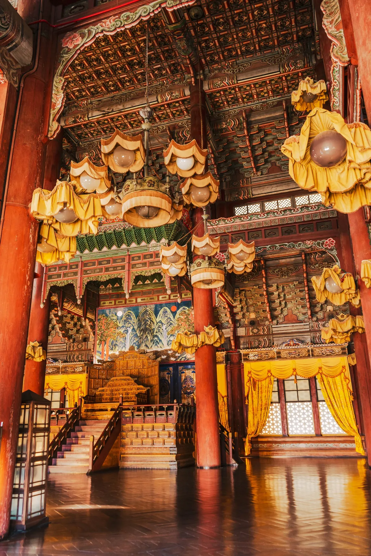 Interior of a palace hall with red pillars, a painted ceiling, yellow silk curtains and hanging lamps, a raised throne and a mountain screen behind, on a polished floor.