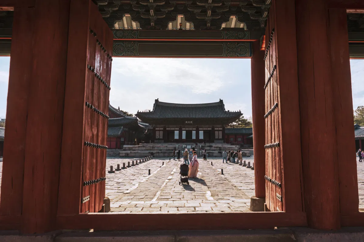 Open red wooden doors framing a palace courtyard with a hall at the far side, a woman in pale pink hanbok pushing a stroller across the stones.