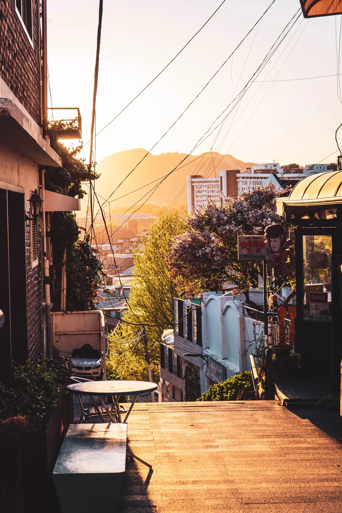 Narrow hillside street in warm low sunlight, with a small cafe table in the shade, a flowering tree and rooftops and a ridge in the golden haze beyond.