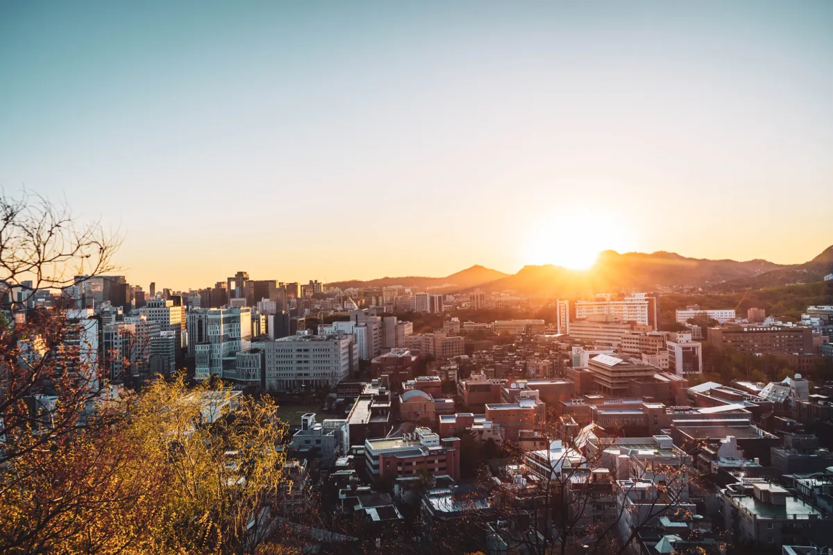 Sun low over the hills above Seoul at dusk, a warm orange glow on the skyline, office towers at left and autumn-coloured branches in the foreground.