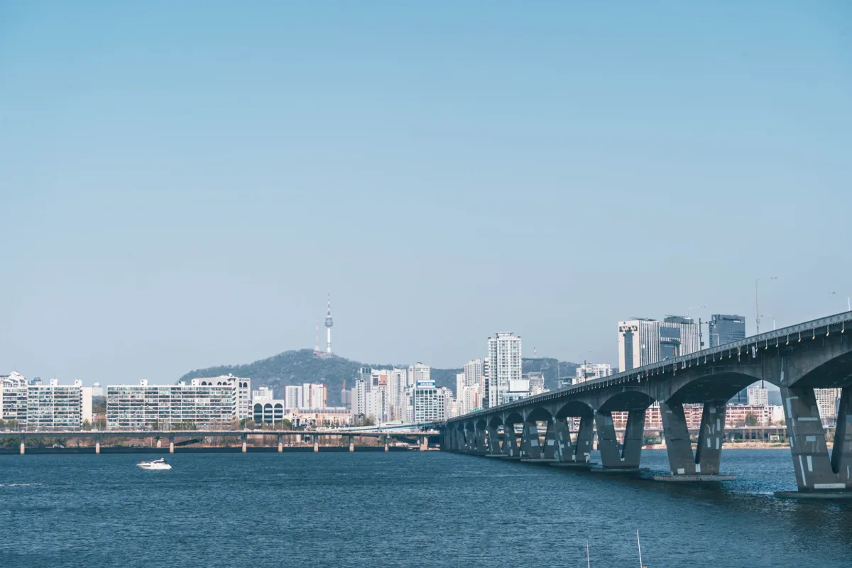 Han River in Seoul with a long concrete bridge at right, a small white boat on the water, and N Seoul Tower on its hill against a clear blue sky.