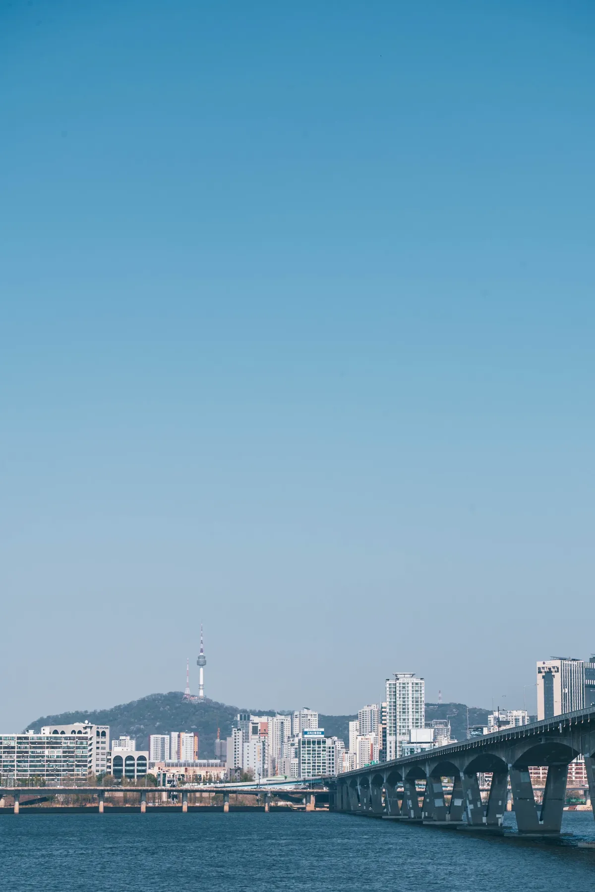 Tall frame of clear blue sky above the Han River skyline, with a long bridge at lower right and N Seoul Tower on its hill in the distance.