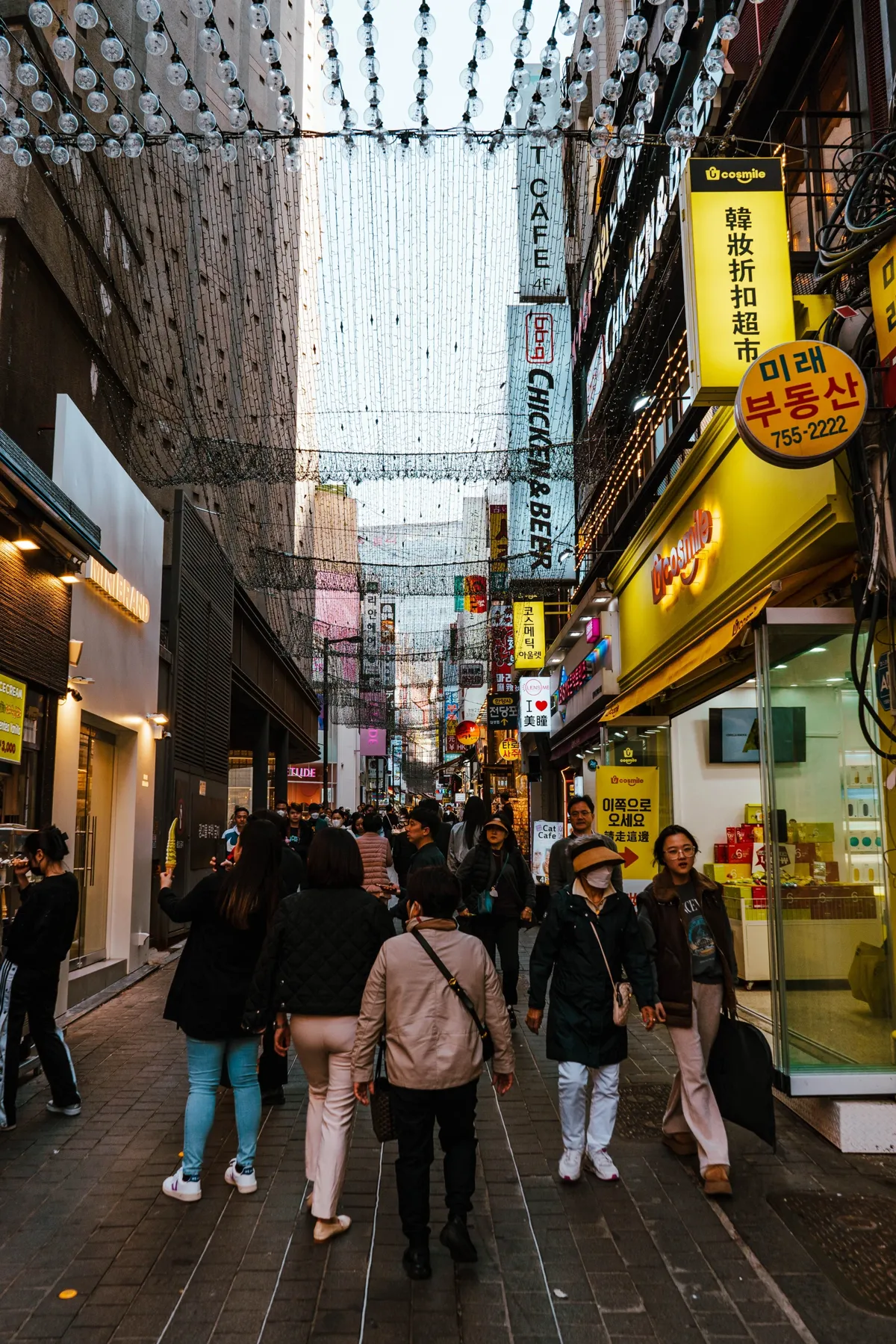 Pedestrian street in Myeongdong crowded with people, strings of lights overhead and yellow and white shop signs in Korean and English on both sides.