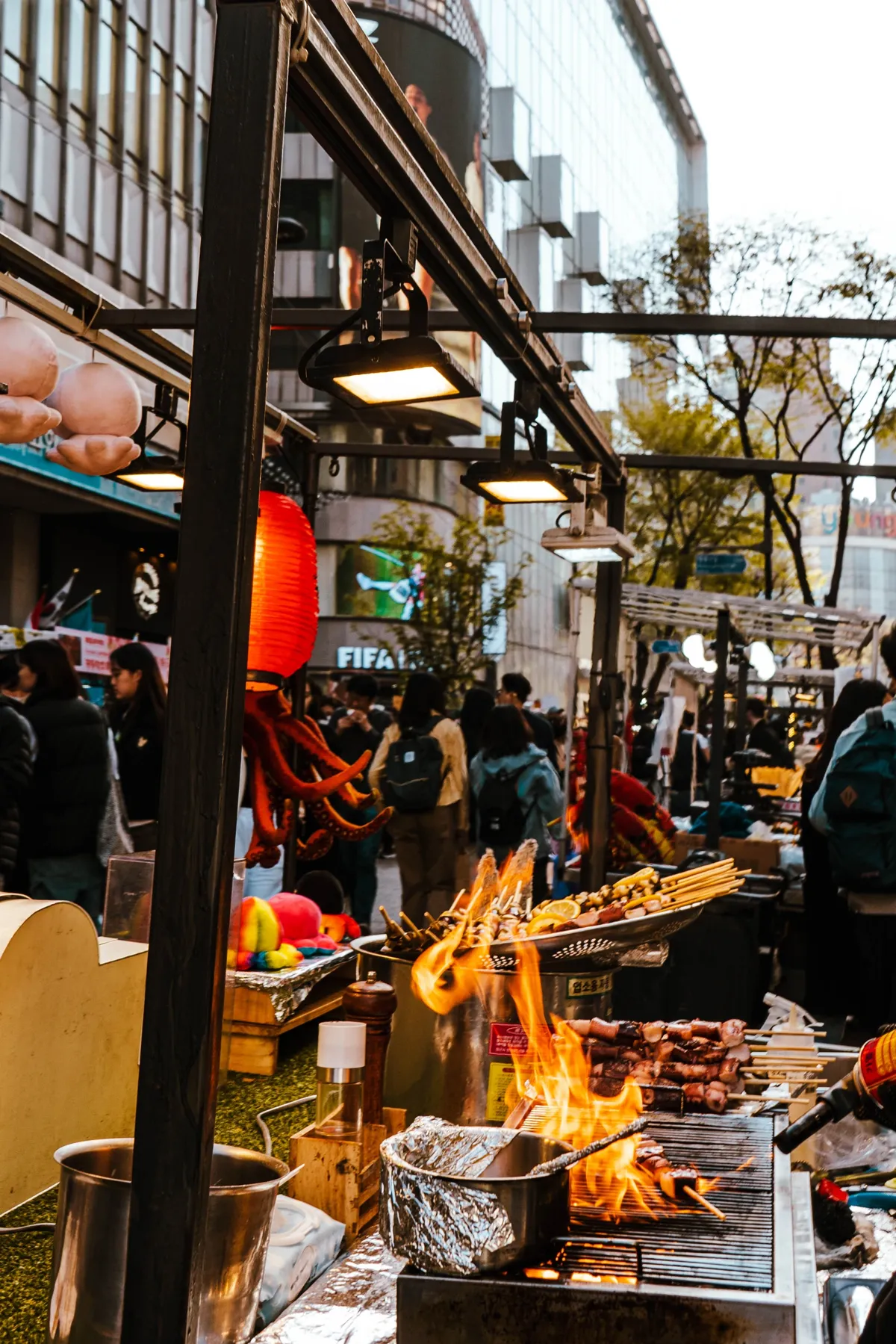 Street food stall with skewers grilling over flames, a red lantern and an orange octopus hanging at left, and pedestrians and buildings behind.