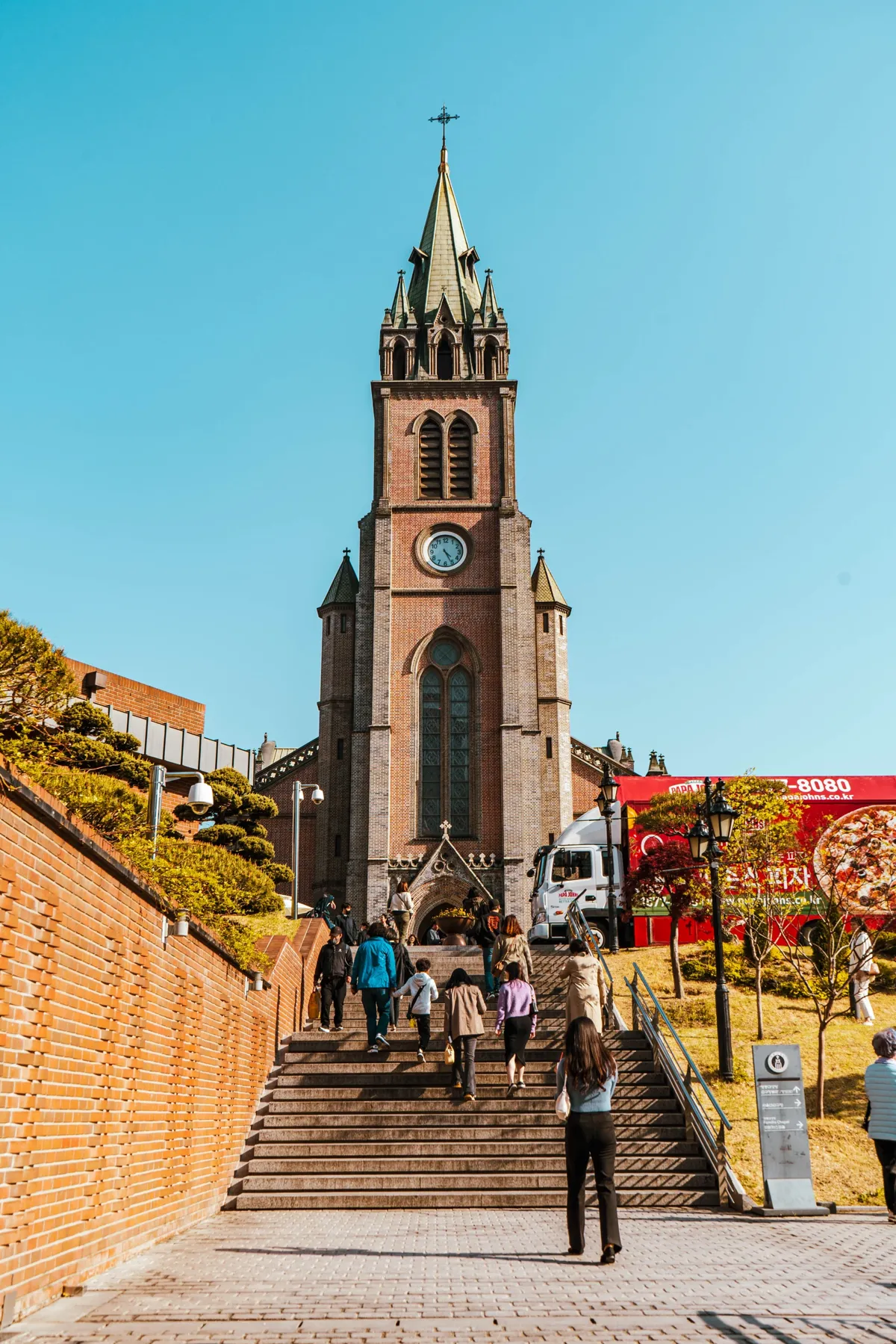 Brick church with a tall clock tower and green spire at the top of a wide stone staircase, visitors climbing, a brick wall at left and a clear blue sky.