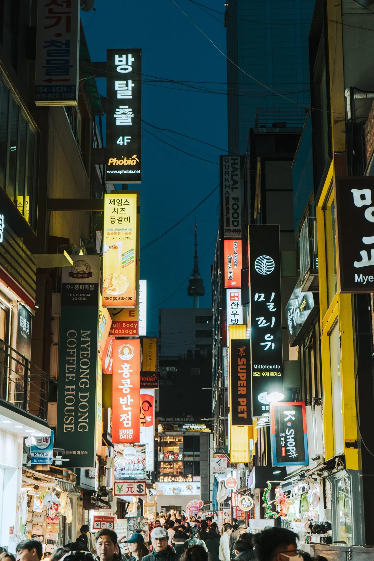 Narrow Myeongdong street at dusk lined with lit signs in Korean and English including a coffee house sign, crowds below and N Seoul Tower against the dark blue sky.