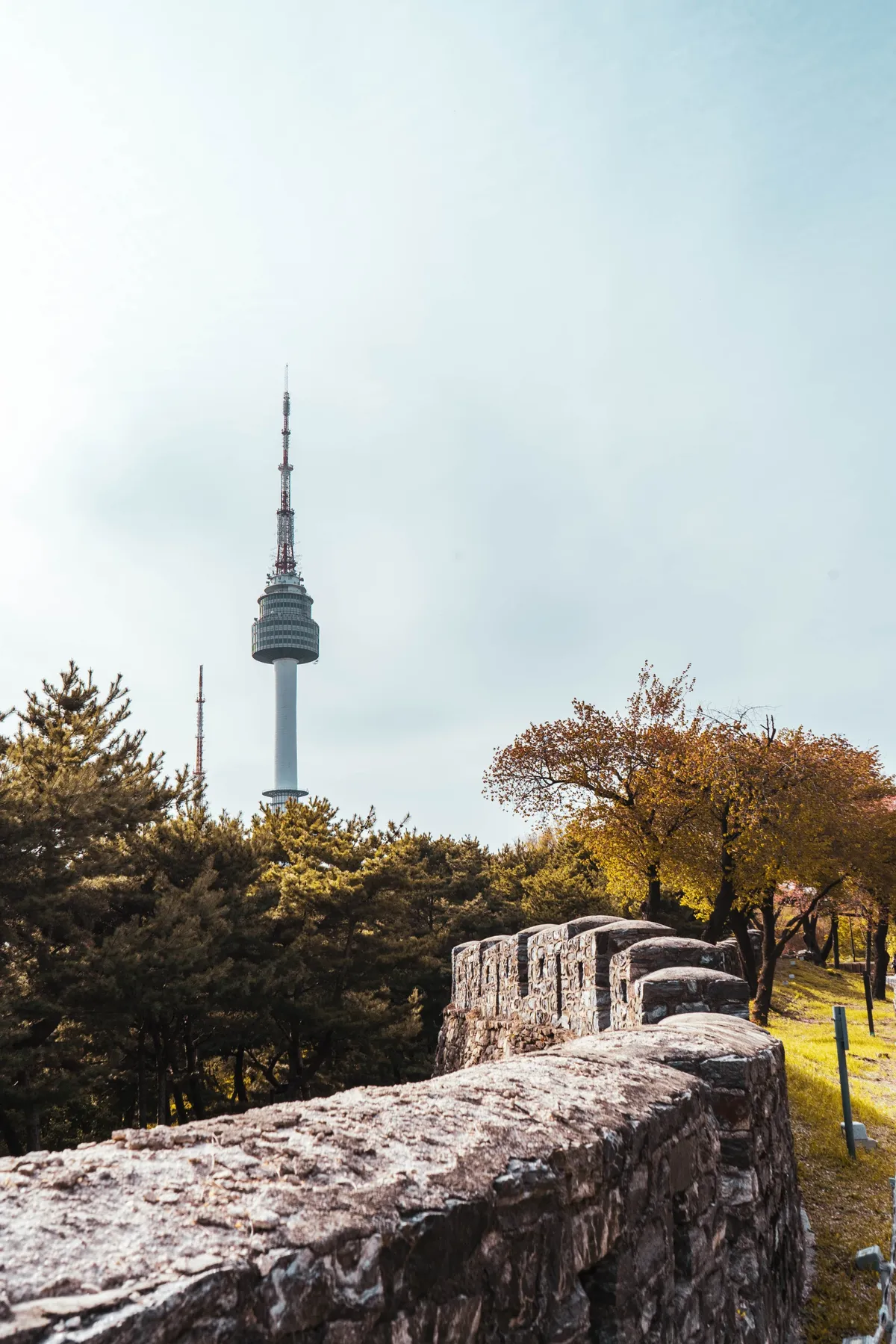 N Seoul Tower rising above pine and autumn trees behind a rough stone fortress wall in the foreground, under a hazy pale sky.