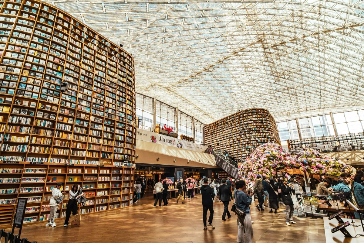 Large atrium library with towering curved bookshelves, a glass roof, visitors walking across a wooden floor and a flower-covered display at right.
