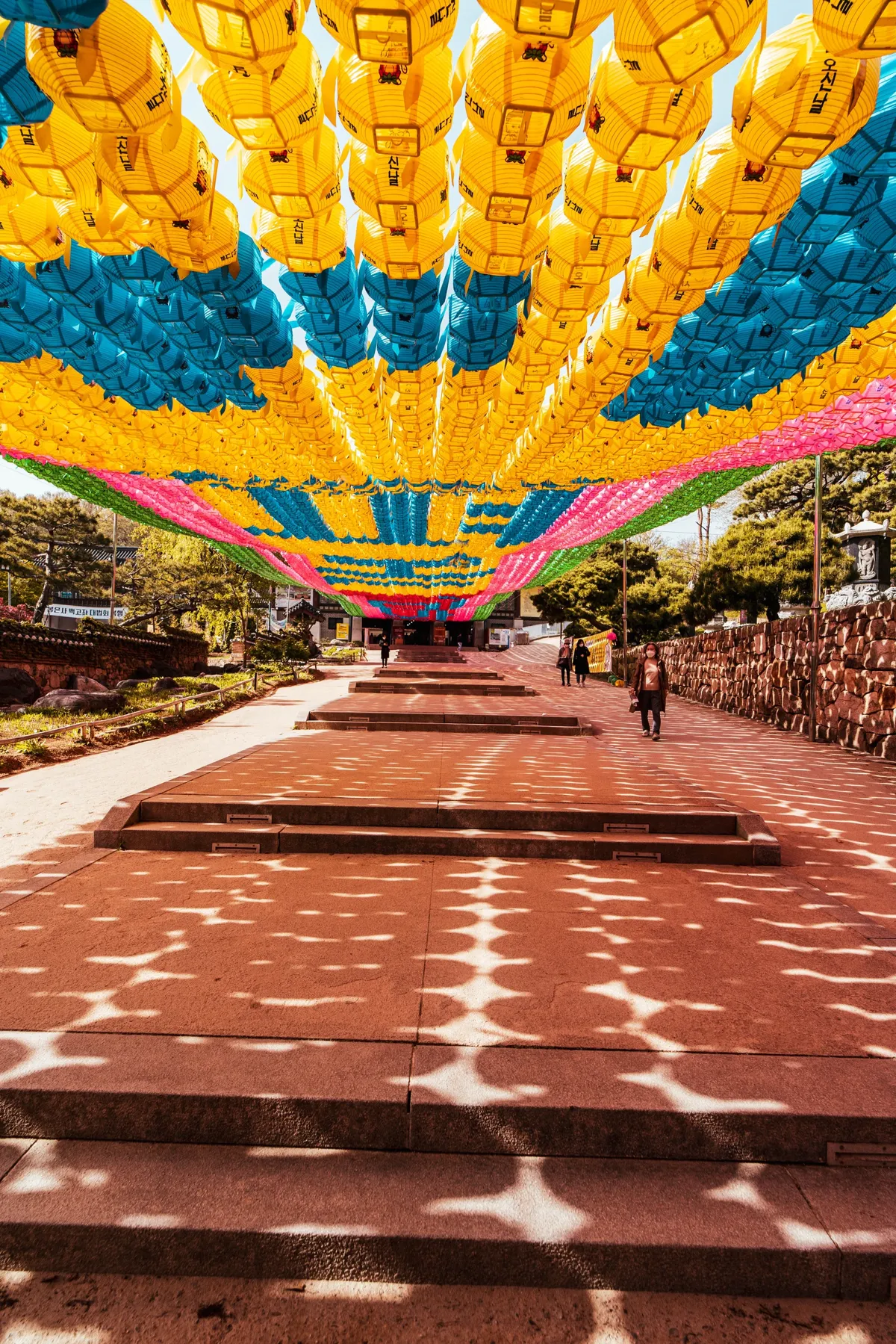 Path under a canopy of yellow, blue and pink paper lanterns at a temple, sunlight casting dappled shadows on red-brown paving, a few people in the distance.