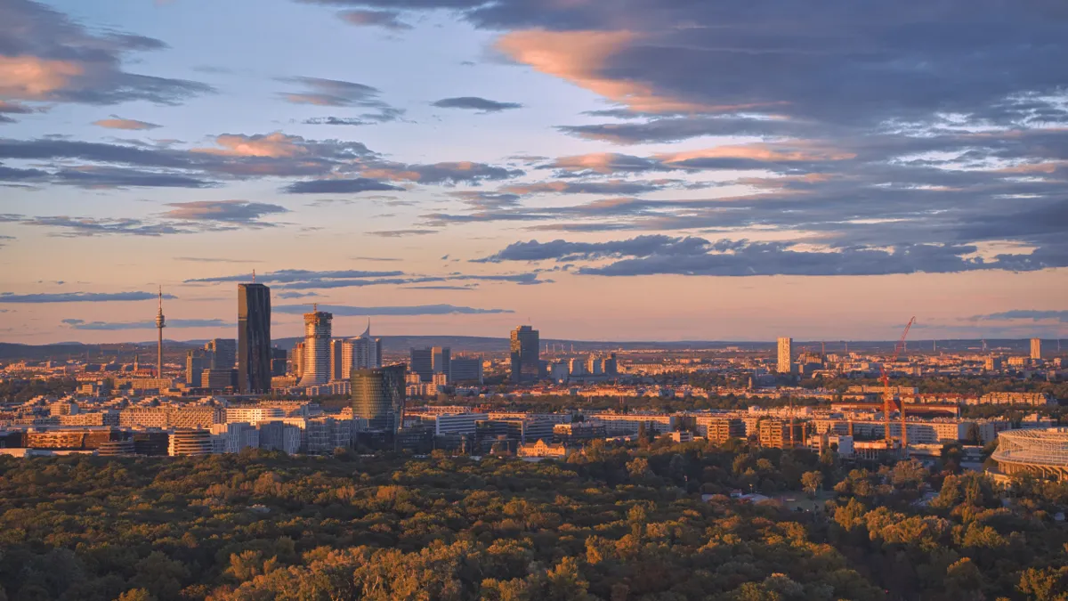 Vienna's skyline of modern towers at golden hour, seen over green woodland under a sky of pink and grey clouds.