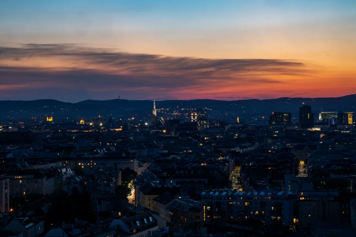 Vienna's rooftops at dusk under an orange and pink sky, with a church spire and lit windows across the city.