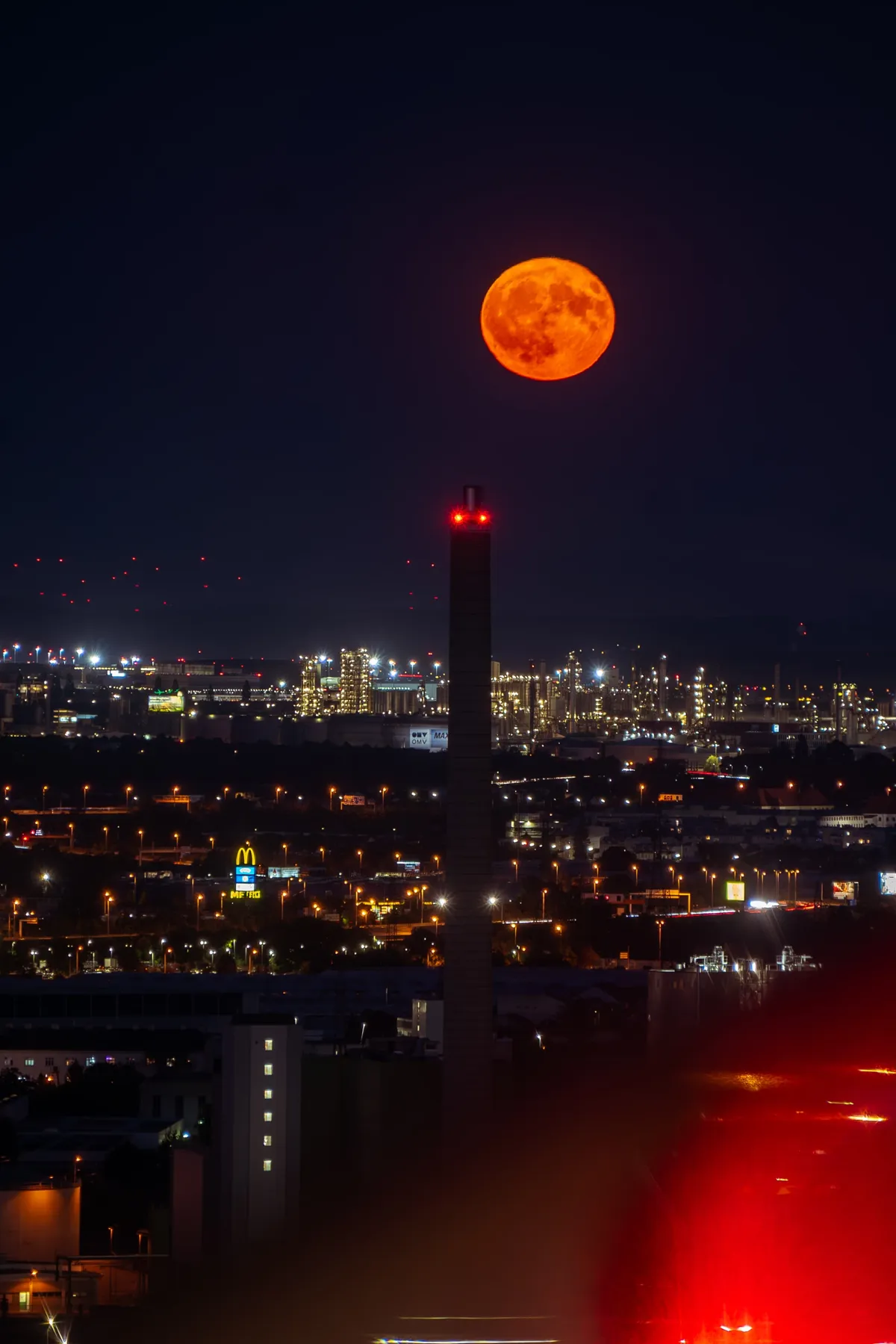 A large orange moon rises over a tall industrial chimney and the lights of Vienna's outskirts at night.