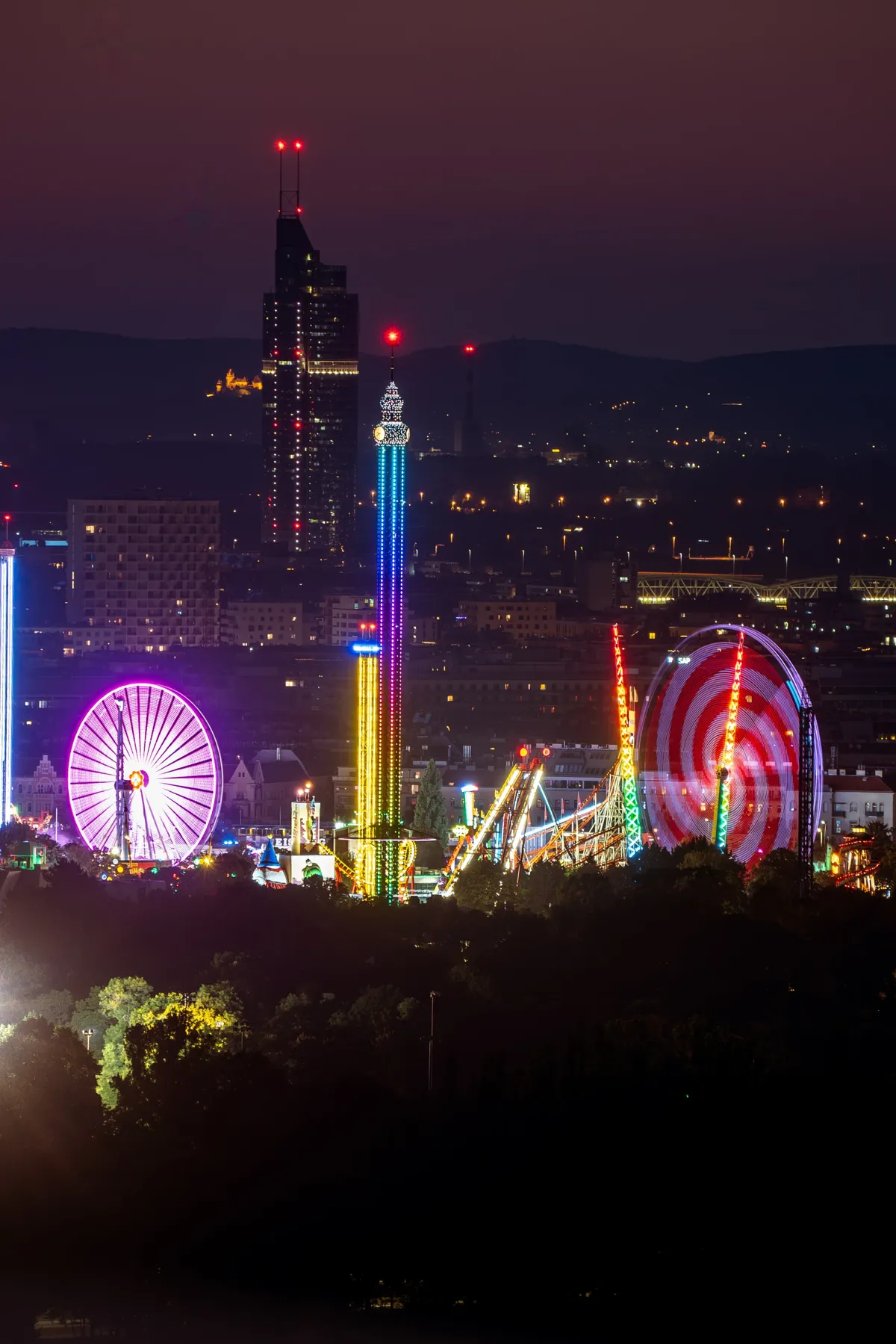 Brightly lit fairground rides, including a purple Ferris wheel and a tall drop tower, glow at night in Vienna.
