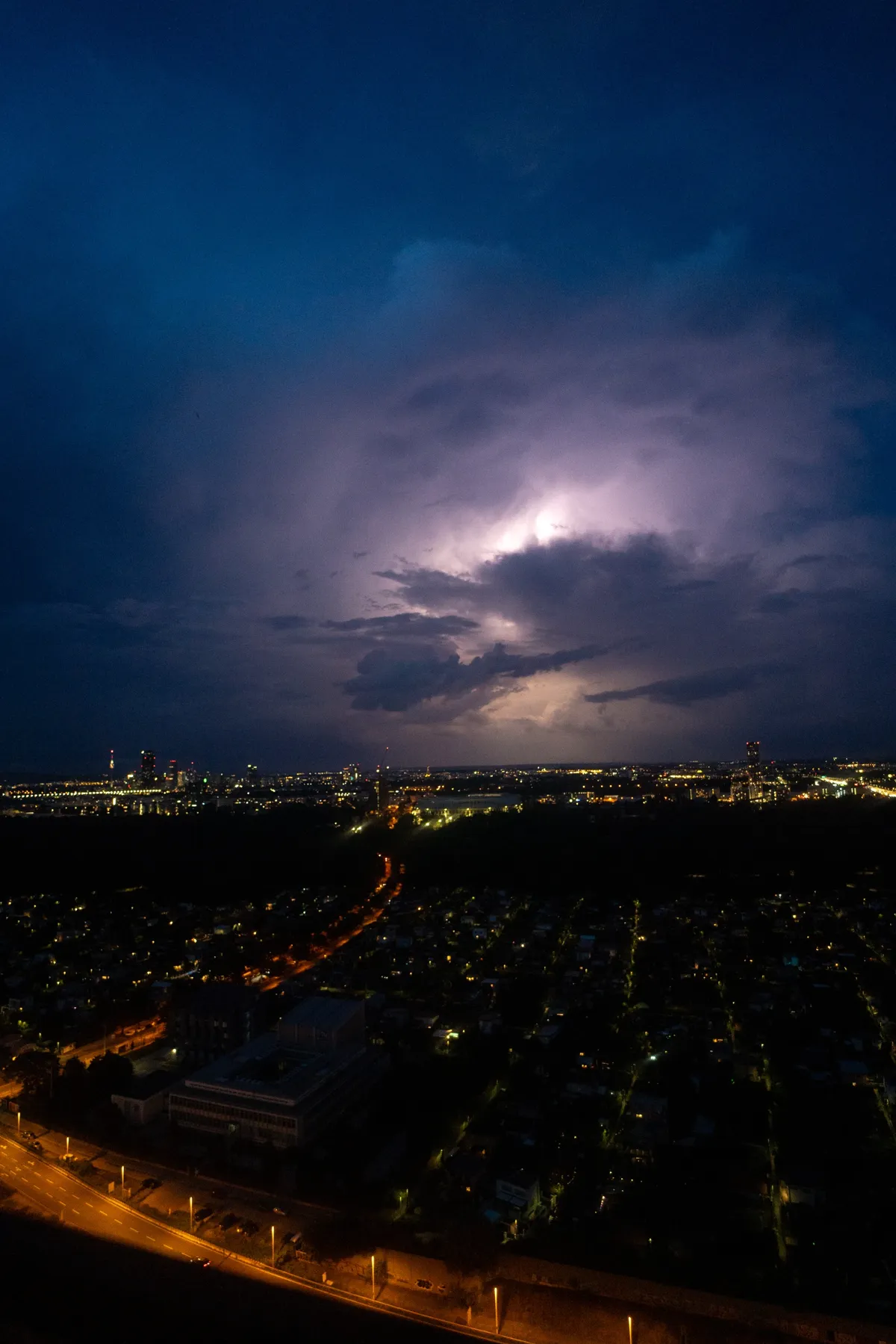 A thunderstorm lights up purple clouds over Vienna at night, seen from above with the city and a road below.
