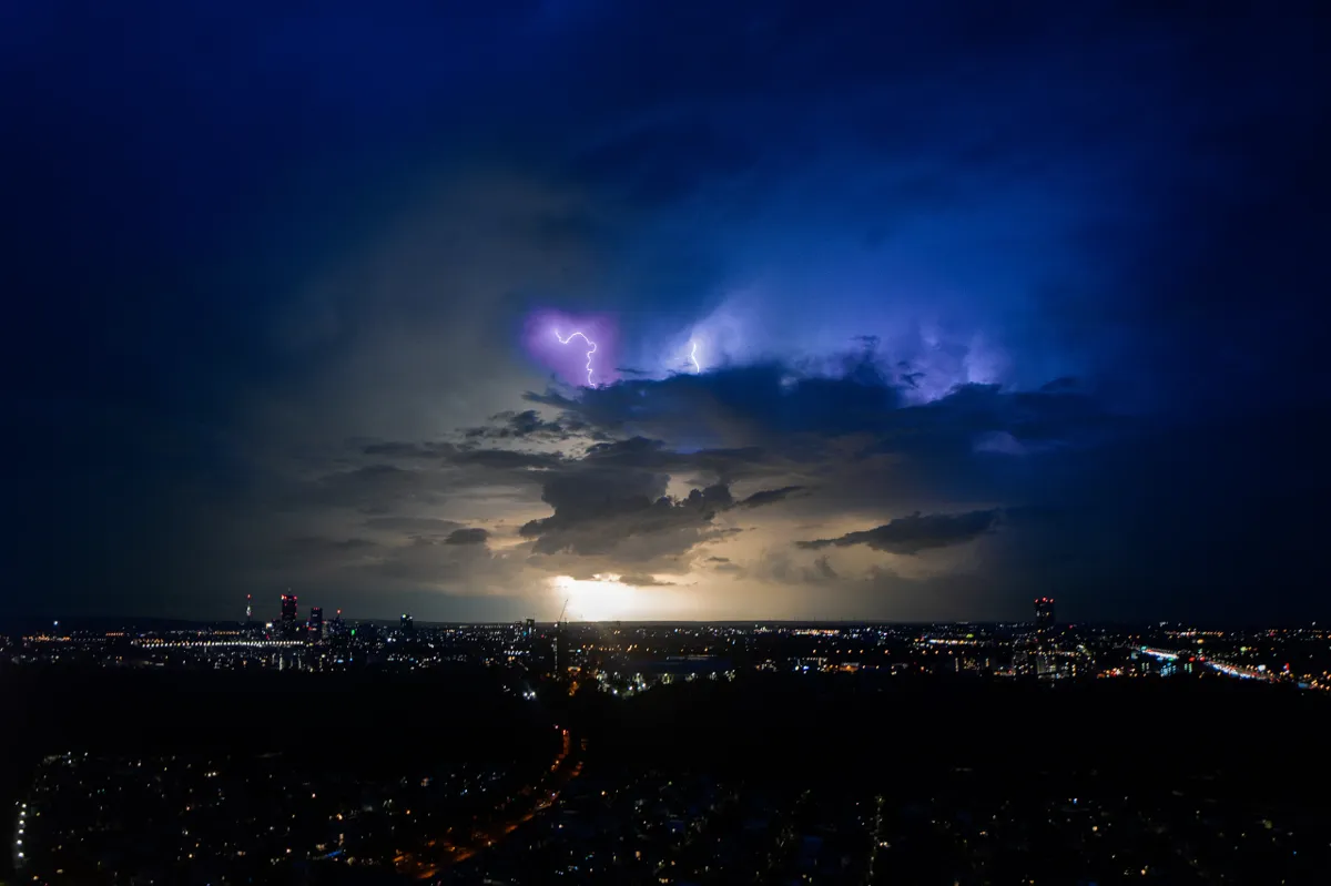 Forked lightning flashes in blue and purple storm clouds above the lights of Vienna at night.