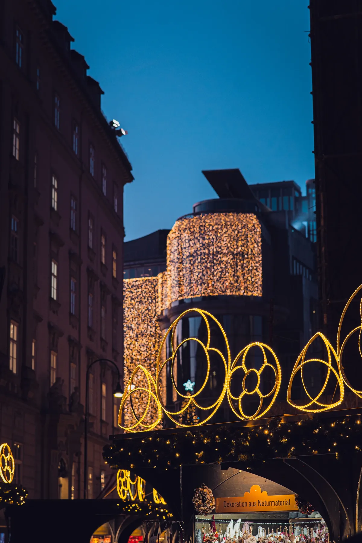 Golden circle-shaped Christmas lights and glowing curtains of lights above a market stall in Vienna at blue hour.