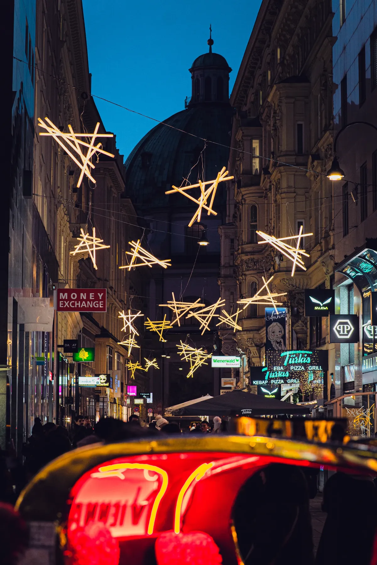 Star-shaped Christmas lights hang over a Vienna shopping street toward a dark church dome, with a red illuminated carriage ahead.