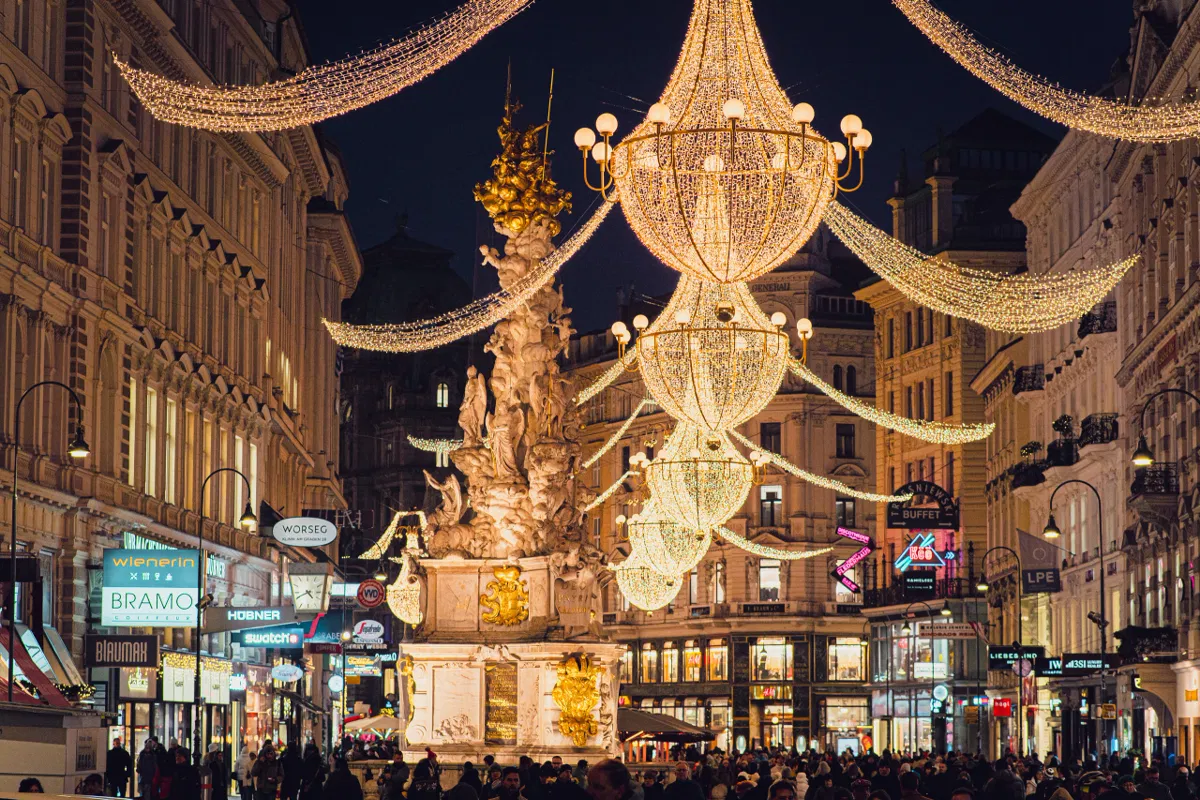 Chandelier-shaped Christmas lights hang above a crowded Vienna street, with an ornate baroque column monument below them.