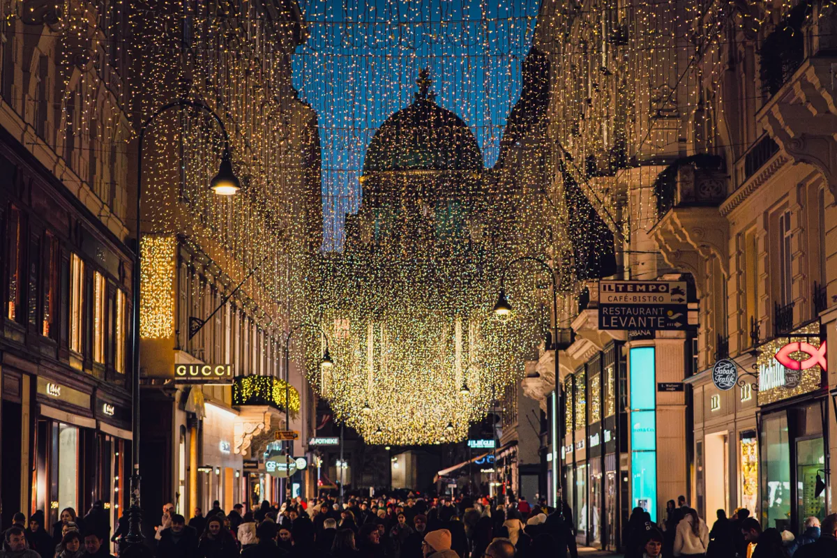 A crowded Vienna shopping street under curtains of fairy lights at dusk, with a green dome visible at the end.