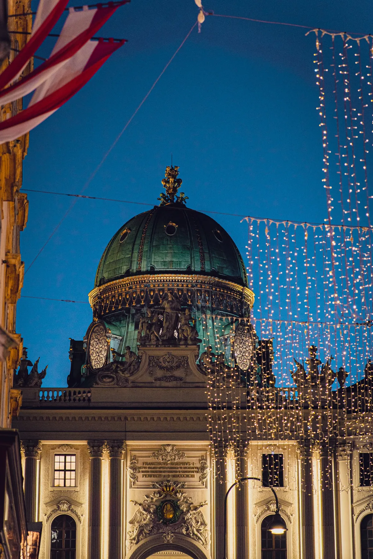 The green copper dome of the Hofburg in Vienna, with red and white flags and hanging lights at dusk.