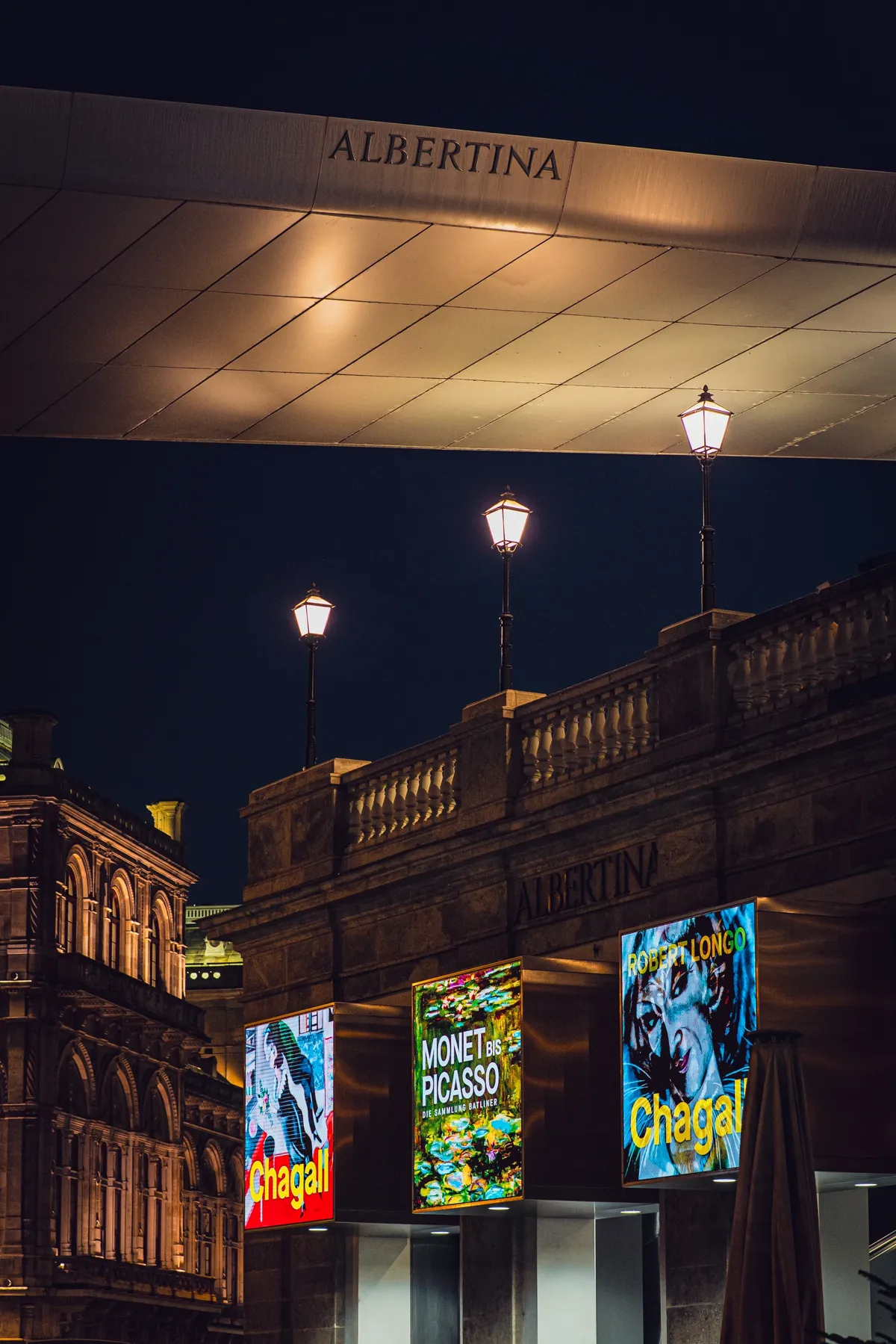A modern museum canopy above street lamps and lit exhibition posters in Vienna at night, beside an old building.