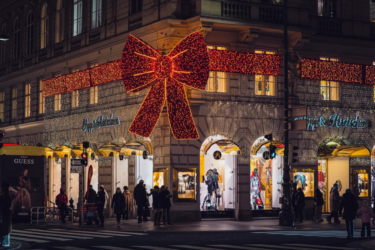 A large red illuminated bow decorates a corner building in Vienna, with lit shop windows below and pedestrians crossing.