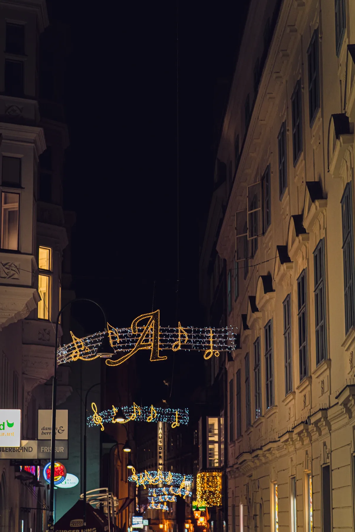 Christmas lights shaped like musical notes hang across a narrow Vienna street at night between tall old buildings.