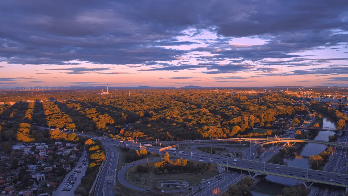 Autumn forest glows gold at sunset above Vienna, with motorways, a river bend and a distant chimney under purple clouds.