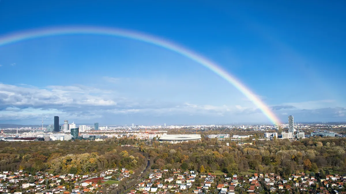 A rainbow arcs across a blue sky over Vienna's skyline, an oval stadium, autumn woods and suburban houses.