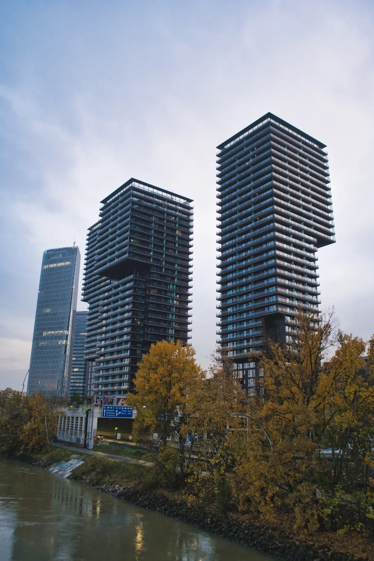Two tall dark residential towers with balconies rise above autumn trees beside a river in Vienna, under a cloudy sky.