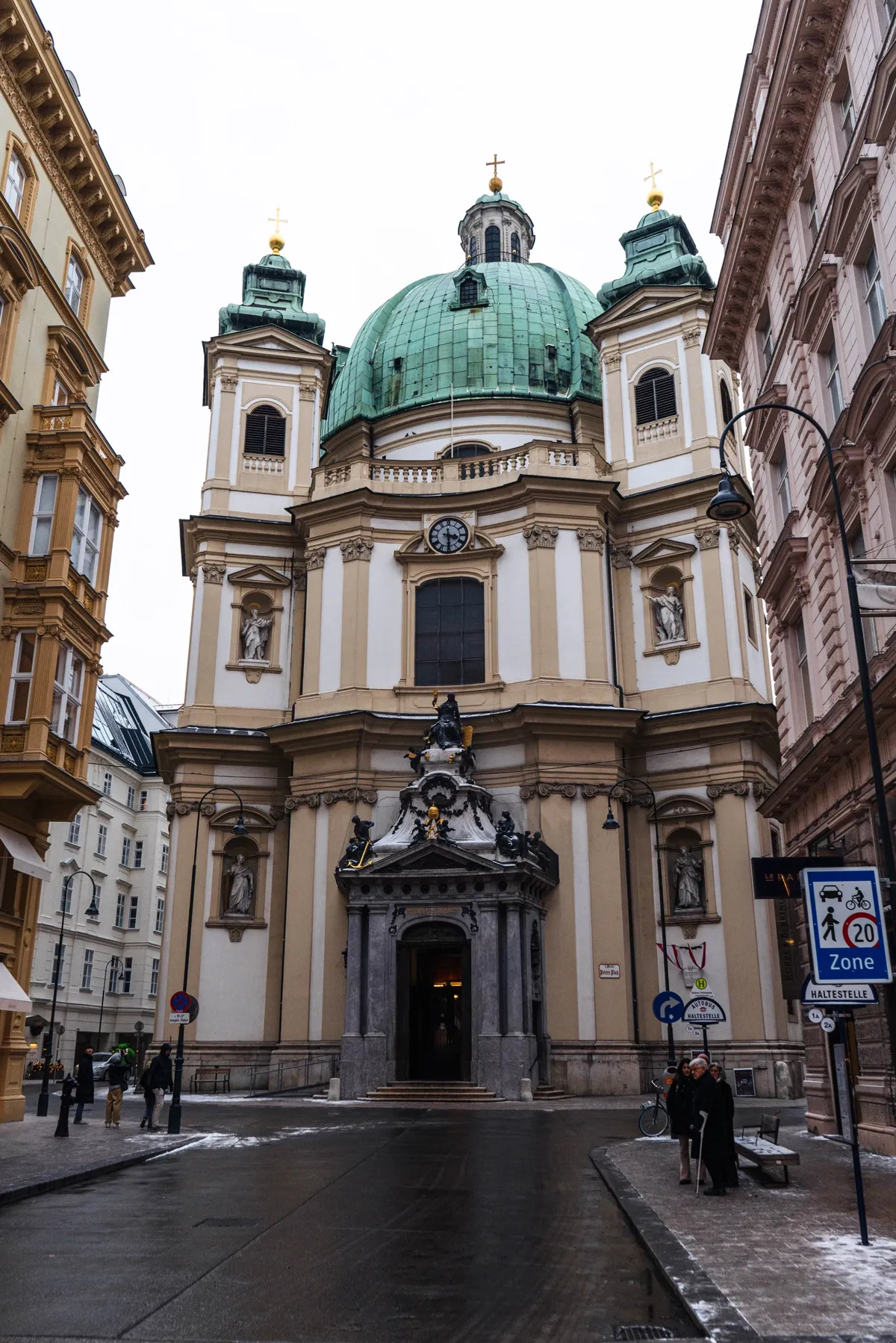 A baroque church with a green dome and twin towers stands on a wet Vienna street between old apartment buildings.