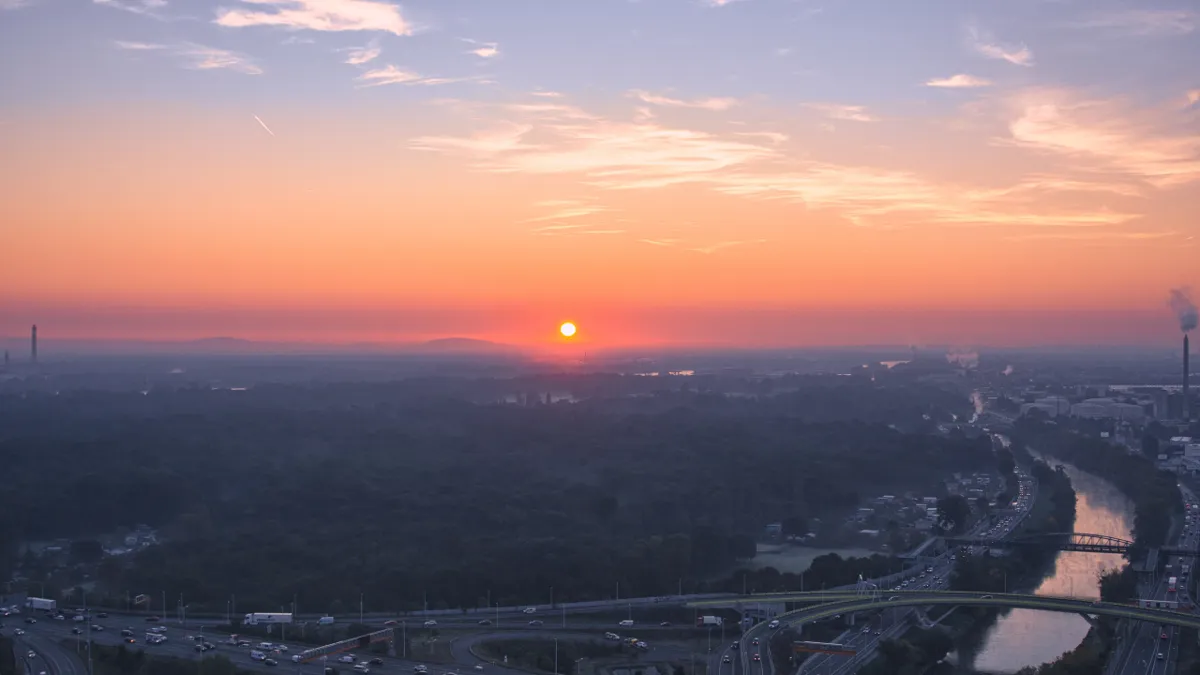 The sun rises as an orange disc over hazy forest and a river beside motorways, seen from above Vienna.