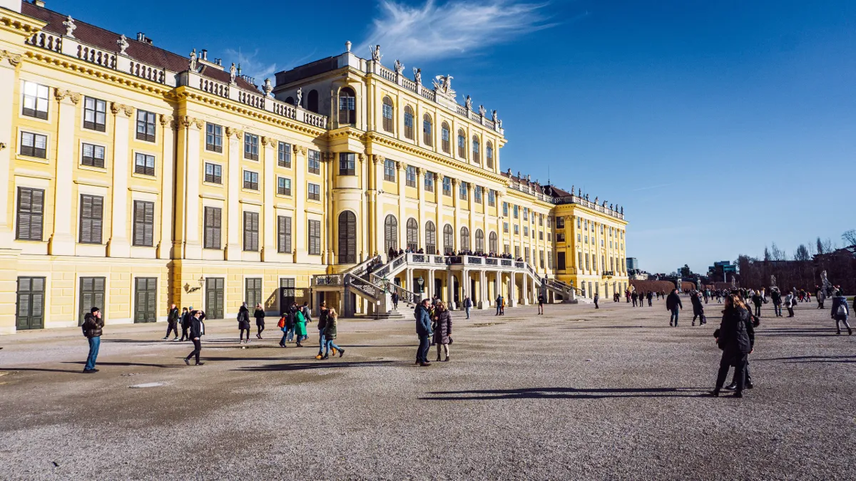 People walk across the gravel forecourt of a large yellow baroque palace in Vienna, under a deep blue sky.